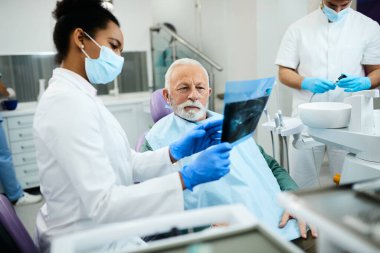 African American dentist showing to senior man his orthopantomogram during dental appointment.