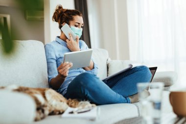 Young businesswoman with protective face mask talking on the phone while using touchpad and going through reports at home. 