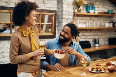 Happy black woman giving her husband strawberries while drinking wine at home. Focus is on man. 
