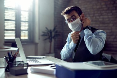 Young businessman putting on protective face mask while working at his office desk during virus epidemic. 