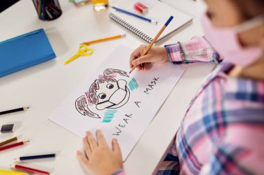 Close-up of creative girl with face mask drawing on the paper while homeschooling during coronavirus pandemic. 