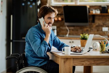 Evdeki cep telefonuyla iletişim kurarken tekerlekli sandalyede çalışan mutlu genç bir işçi.. 