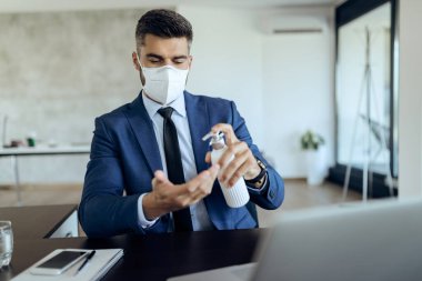 Businessman with face mask disinfecting hands while working at office desk. 