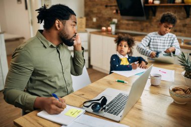 Black father talking on the phone and taking notes while working at home and babysitting his kids. 