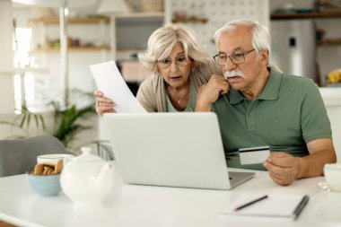 Worried mature couple using laptop and credit card while checking their online bank account and analyzing their finances at home. 