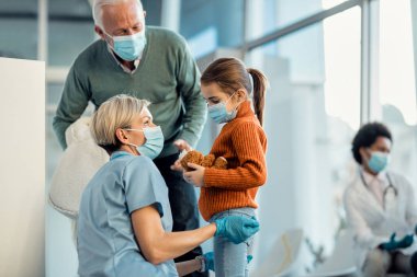Sad little girl holding her plush toy while talking to a nurse in the hospital during COVID-19 pandemic.