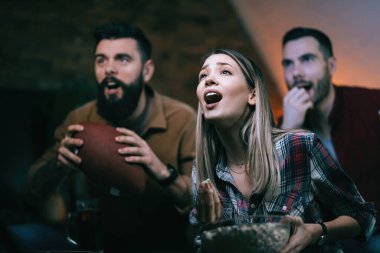 Small group of young people shouting while cheering for their team and watching sports match on TV. Focus is on woman. 