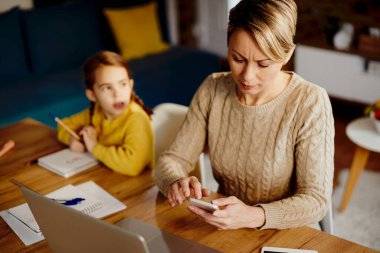 Female entrepreneur working and text messaging on mobile phone while being with her daughter at home. 