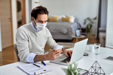 Male entrepreneur wearing protective face mask and texting on mobile phone while working on a computer at home. 