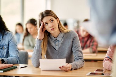 Displeased college student holding her test results and listening to a professor during a class in the classroom.