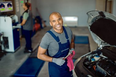 Happy African American auto repairman working in a workshop and looking at camera. 