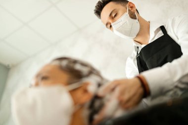 Low angle view of hairstylist with protective face mask washing customer's hair at the salon during coronavirus epidemic. 