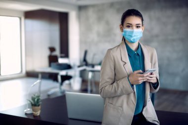 Young businesswoman wearing protective face mask while using smart phone in the office and looking at camera. 