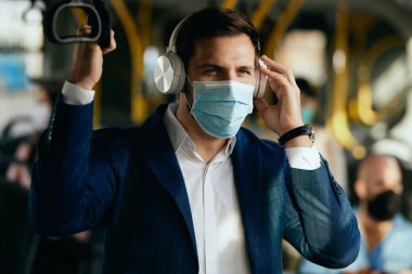Businessman wearing protective face mask while listening music over headphones and commuting to work by bus. 