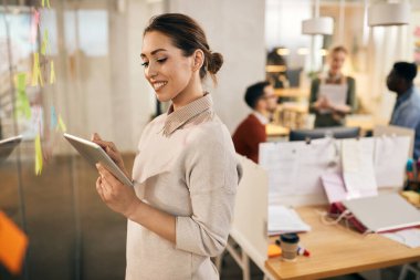 Happy creative woman using digital table while standing in front of mind map on glass wall in the office. Her colleagues are in the background. 
