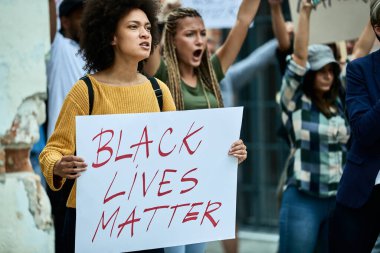 African American woman carrying Black Lives Matter banner protesting with crowd of people on the streets. 