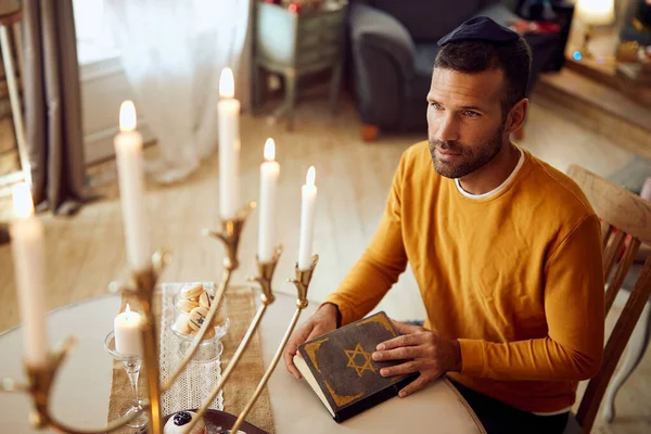 Jewish man reading Tanakh and looking at burning candles in menorah while celebrating Hanukkah at home.