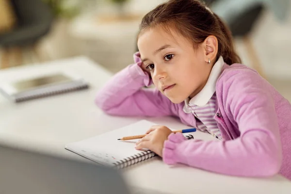 Small girl studying at home and writing in notebook while leaning on a desk.