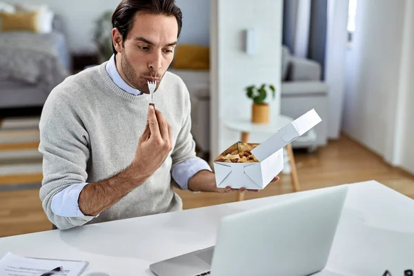 Young businessman surfing the net on a computer while eating at home.