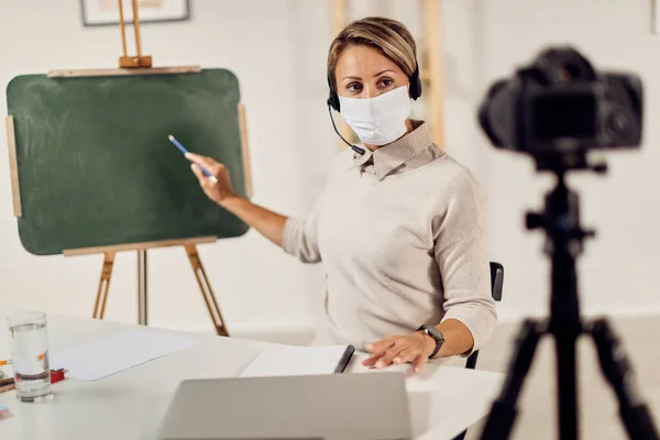 Professor with protective face mask pointing at chalkboard while teaching her students during live stream due to coronavirus pandemic.