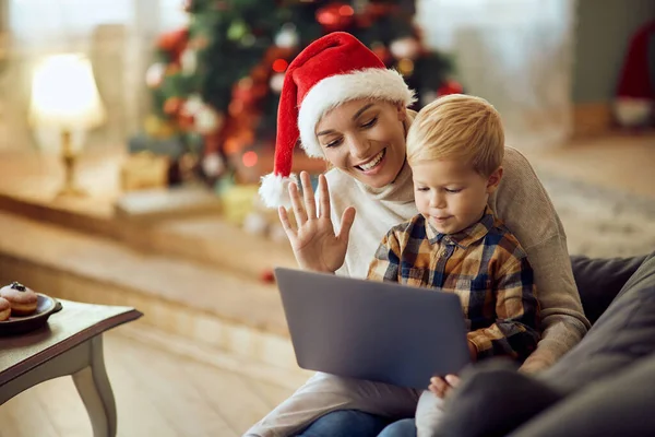 Happy mother and her small boy using laptop and greeting someone while making video call during Christmas day at home.