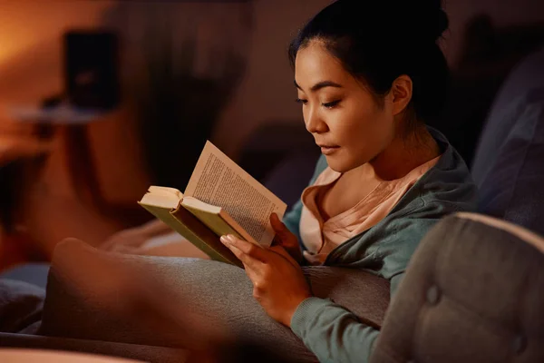 Young Asian woman relaxing on the sofa and reading a book in the evening.