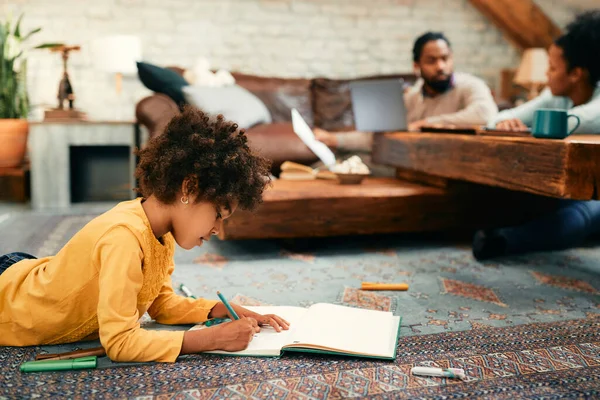 African American little girl enjoying in coloring on paper while her parents are working in the background at home.