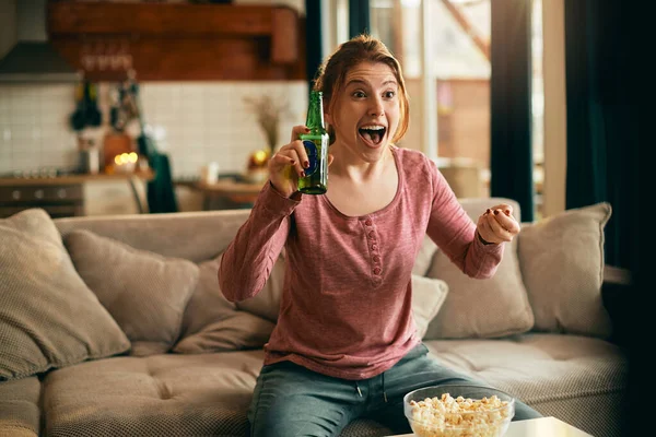 Excited woman watching sports match on TV while drinking beer and eating popcorn in the living room.