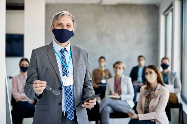 Happy business leader with digital tablet standing in board room while holding seminar to group of coworkers during COVID-19 pandemic.