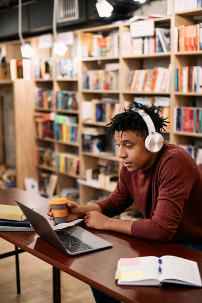Young black student using laptop while drinking coffee and studying at university library.
