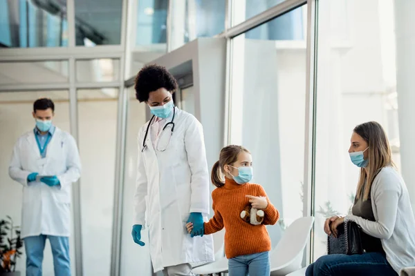 African American doctor holding hands with a small girl and taking her away from mother to medical exam in the hospital. They are wearing face masks due to coronavirus pandemic.