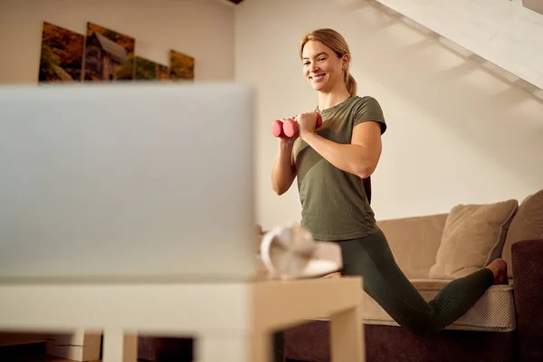 Low angle view of happy athletic woman using laptop while practicing with hand weights n the living room.