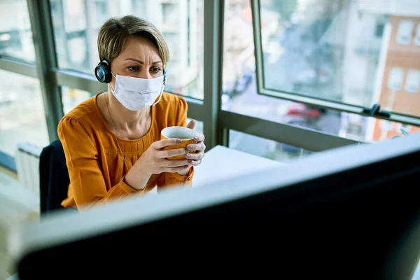 Businesswoman with face mask holding a cup of coffee while reading problematic e-mail on desktop PC in the office.