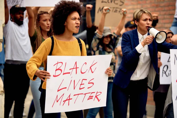 Large group of protesters marching at demonstrations against police brutality. Focus is on African American woman holding Black Lives Matter placard.