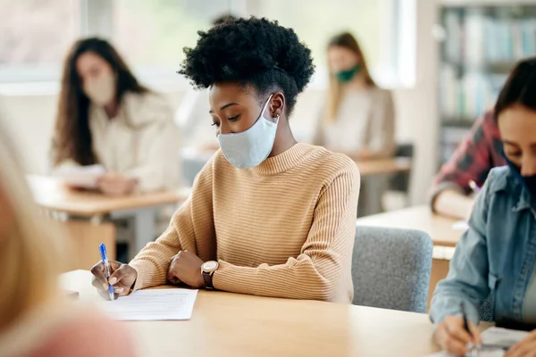 African American female student taking notes and wearing protective face mask during a lecture in the classroom.