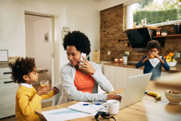 Happy working black mother using mobile phone and laptop while communicating with her daughter at home.