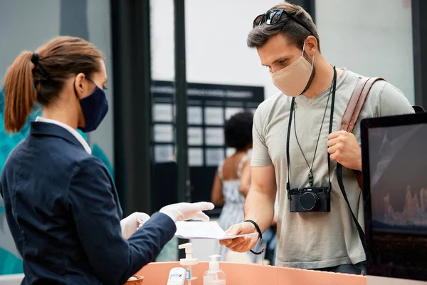 Man with face mask reading application form and talking to receptionist while checking in a hotel.