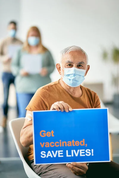 Mature man with face mask inviting people to get vaccinated against coronavirus and save lives while holding placard at vaccination center.