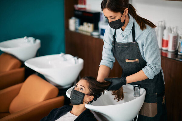 Hairdresser washing hair of female client during appointment at hair salon. Both of them are wearing protective face masks.