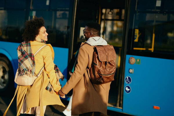 Happy African American couple talking while holding hands and walking toward a bus on the station.