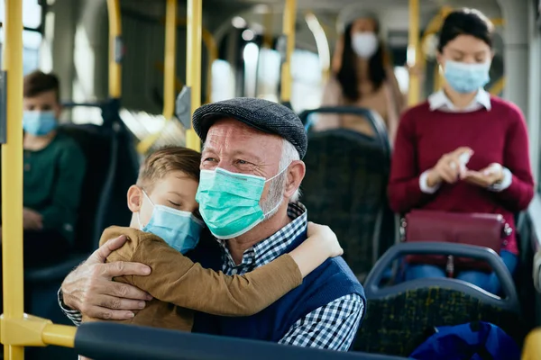 Caring grandfather holding his sleepy grandson while traveling by public transport and wearing protective face masks.