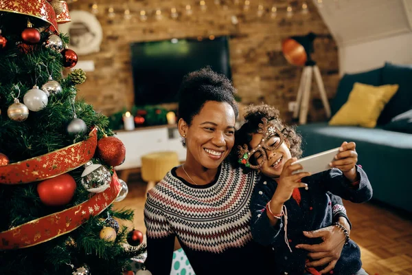 Happy African American mother and daughter taking selfie with smart phone and having fun on Christmas at home.