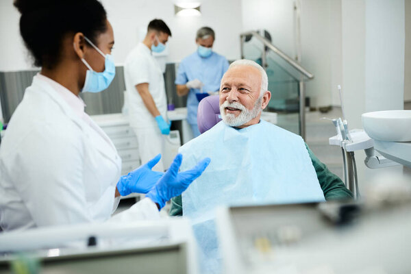 Black female dentist communicating with mature man during teeth exam at dentist's office. Focus is on man. 