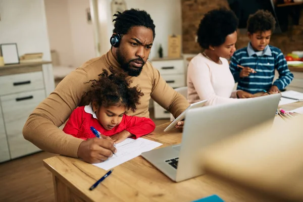 African American working father taking notes while using touchpad and laptop at home. 