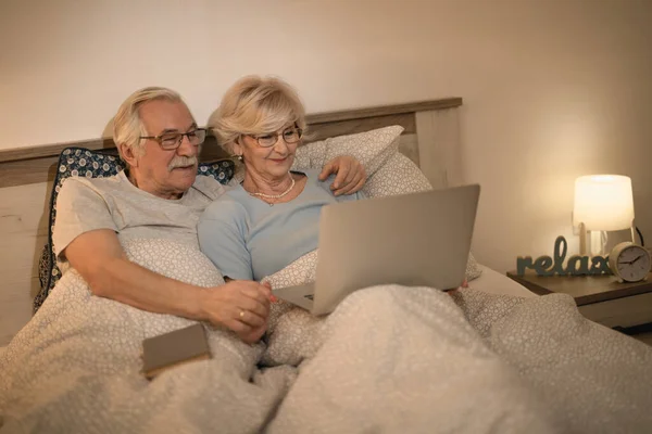 Smiling senior couple relaxing in bedroom and surfing the Internet on laptop.