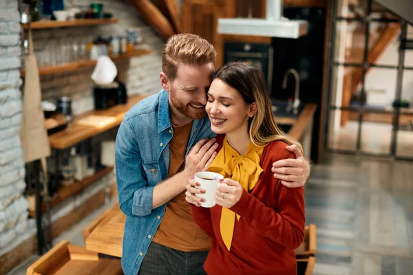 Happy redhead man embracing his girlfriend who is having a cup of coffee at home.