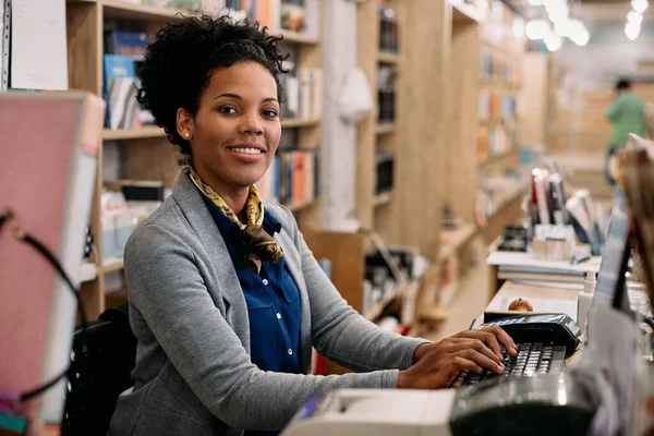 Happy African American woman working on a computer at bookstore and looking at camera.