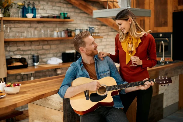 Happy woman drinking wine and talking to her boyfriend who is playing acoustic guitar at home.