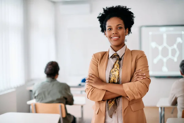 Happy black female professor standing with arms crossed in the classroom and looking at camera.