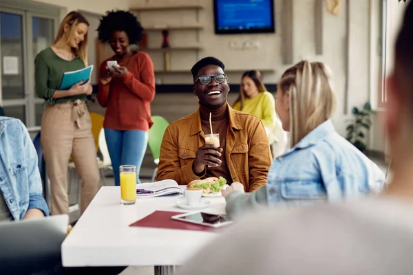 African American university student laughing while communicating with female colleague on lunch break in cafeteria.